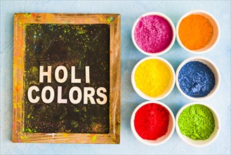 Color powder white bowls near wooden slate with holi color text