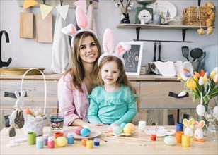 Happy woman sitting with daughter bunny ears near easter eggs