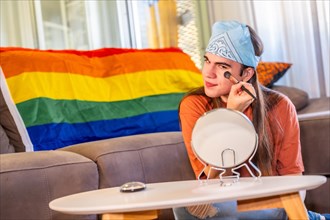 Transgender young man making up using brush and mirror sitting in the living room next to a rainbow lgbt flag at home