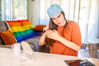 Young transgender person combing using a mirror in the living room next to a sofa with a lgbt rainbow flag