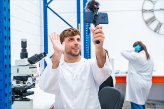 Young handsome scientist waving while broadcasting using phone in a research laboratory
