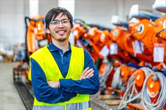 Proud japanese chief engineer of a robotic industry standing with arms crossed next to the assembly line