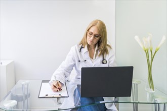Female doctor working on laptop and writing on clipboard in medical office