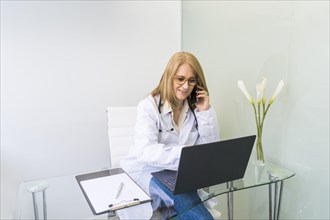 Smiling female doctor talking on the phone to a patient while looking at her medical records on laptop in medical office
