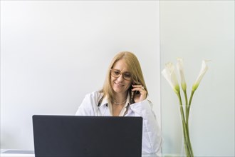 Female doctor talking on the phone to a patient while looking at her medical records on laptop in medical office. Copy space