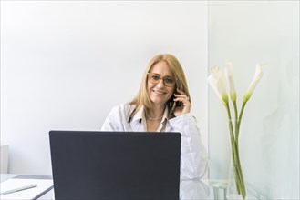 Smiling female doctor talking on the phone to a patient while looking at camera in medical office