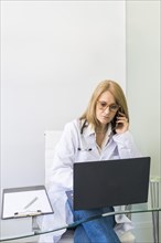 Female doctor talking on the phone to a patient while looking at her medical records on laptop in medical office