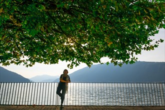 Woman Standing Below a Beautiful Tree with Branch on the Waterfront with Railing to Lake Lugano with Mountain in a Sunny Summer Day in Bissone
