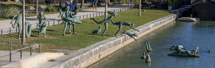 Panoramic photo of a group of bronze sculptures of playful children La Ribambelle Joyeuse by Belgian artist Tom Franzten on the banks and in the Canal du Trevois