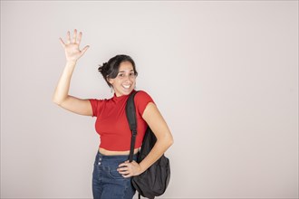 Brunette Latin woman smiling wearing a red t-shirt holding a backpack raising one arm waving on white background. Travel