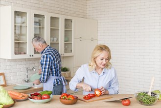 Smiling senior woman cutting red bell pepper with knife looking digital tablet his husband washing dishes kitchen sink