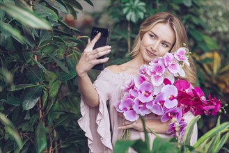 Smiling young woman holding beautiful orchid branches hand taking selfie smart phone