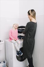 Woman with laundry looking her cute daughter sitting washing machine
