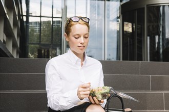 Young businesswoman sitting outside office eating lunch box
