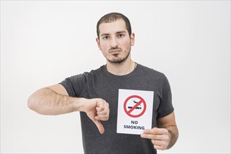 Young man holding no smoking sign showing thumbs down against white background