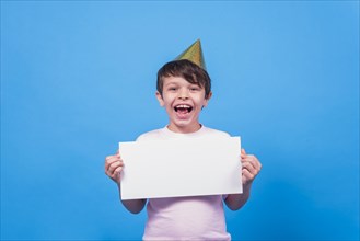 Excited little boy wearing party hat holding blank card hand blue surface
