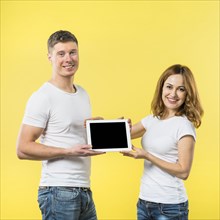 Portrait two happy young couple showing black screen digital tablet against yellow backdrop