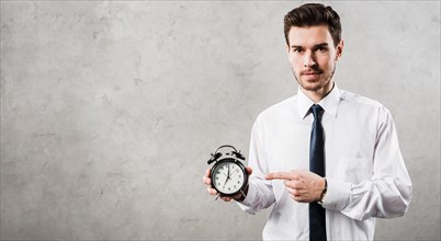 Portrait young businessman pointing black alarm clock standing against grey concrete wall