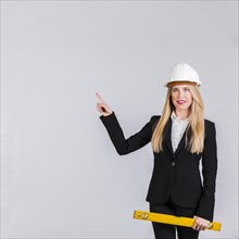 Portrait young female architect wearing hardhat pointing her finger against grey background