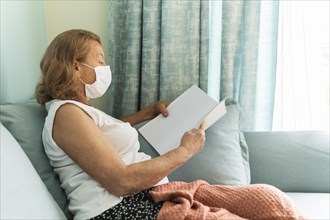 Side view elder woman with medical mask home during pandemic reading book