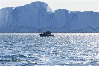 A small excursion boat sails in front of a huge iceberg in the UNESCO World Heritage Ilulissat Icefjord. Disko Bay