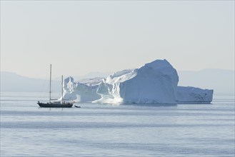 A small sailing ship passes an iceberg in the evening sun. Disko Bay