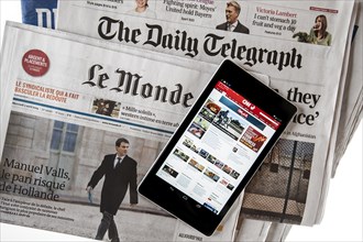 Touchscreen digital tablet showing CNN International online news on top of British The Daily Telegraph and French Le Monde newspapers on white background