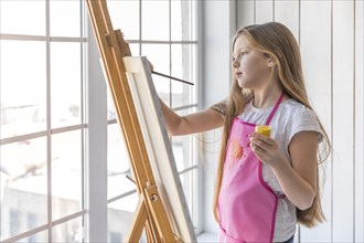 Side view girl holding yellow paint bottle hand painting easel with paintbrush