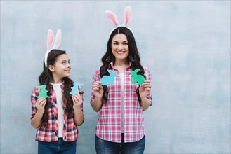Smiling portrait mother daughter holding paper cutout bunny against blue wall