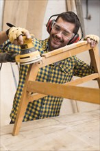 Smiling male carpenter softening edges wooden furniture with sander