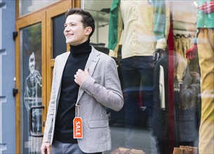Stylish young man wearing jacket with sale tag standing outside shop