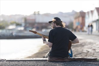A man hipster playing guitar at the waterfront. Mid shot