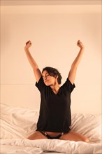 Vertical photo of a woman stretching arms sitting on the bed in the morning