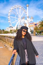 Portrait of a fashionable latin woman in winter clothes standing on a promenade next to the beach in the city