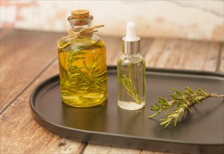 Glass jar with rosemary oil with rosemary branch inside on a brown tray on a wooden table