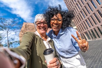 Frontal photo of two multi-ethnic smiling businesswomen taking selfie and gesturing victory with fingers outdoors