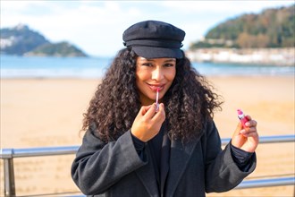 Latin woman making up with lipstick next to the beach during a sunny day in winter