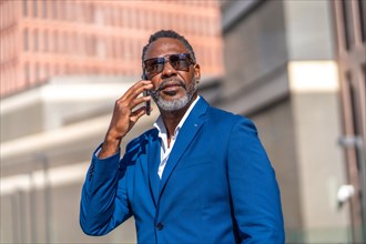 Portrait of a cool african businessman wearing sunglasses ans suit using phone outside a financial building