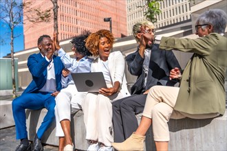 Excited business people celebrating news while using laptop sitting on an urban park bench
