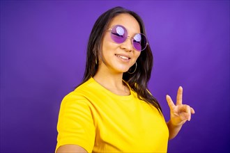 Studio photo with purple background of a hippy chinese woman with sunglasses gesturing peace with fingers