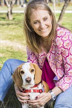 Portrait of smiling woman looking at camera and hugging a Beagle dog while sitting on green grass at the park