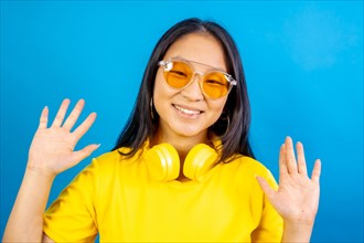 Studio photo with blue background of a happy chinese woman with headphones on neck wearing sunglasses greeting raising hands