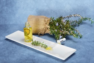 Dropper with rosemary essential oil with fresh branches inside on a white tray next to a burlap bag with flowering rosemary branches