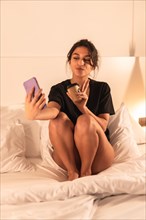 Vertical photo of a streamer taking a selfie from the hotel bed while drinking a coffee