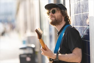 A smiling man hipster wearing sunglasses standing by the wall and playing guitar. Mid shot