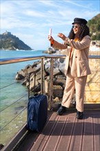 Chic latin woman taking a photo to the sea landscape standing next to luggage in a promenade
