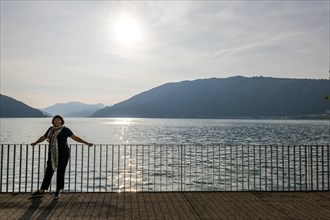 Woman Standing on the Waterfront with Railing to Lake Lugano with Mountain in a Sunny Summer Day in Bissone