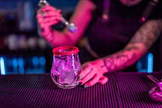 Close-up of a bartender preparing a margarita cocktail adding ice in the counter of a bar with neon lights