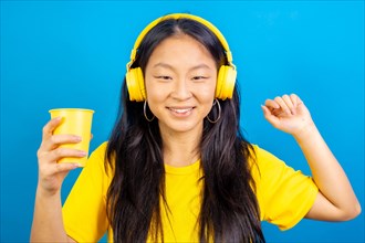 Studio photo with blue background of chinese woman listening to music with headphones and holding a plastic glass