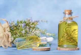 Glass bottle and dropper with rosemary essential oil with fresh branches inside next to a raffia bag with freshly cut flowering rosemary branches on a blue background